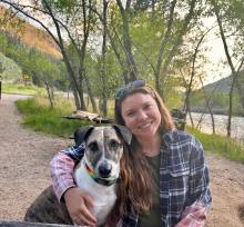 A woman sits near a river with her arm around a dog